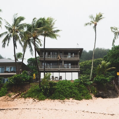 A beach house surrounded by trees and vegetation, with sand in the foreground.